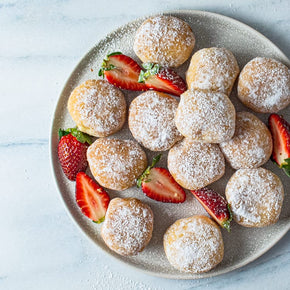 A large round plate with 12 mini donuts with flavoured fillings like Nutella, chocolate, caramel, or berry, dusted with icing sugar and garnished with strawberries on a marble table.