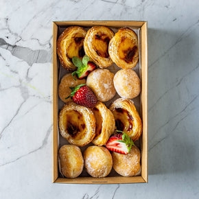 A rectangular cardboard box of 12 mixed Portugese tarts and mini sweet filled donuts garnished with strawberries on a marble table.