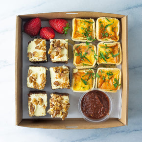 A square cardboard box filled with mini quiche and tomato relish along with serves of sweet slice and fresh strawberries on a marble table.