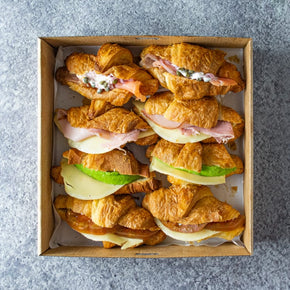 A square coardboard box with eight mini croissants in various fillings like ham, avocado and smoked salmon, on a marble table.
