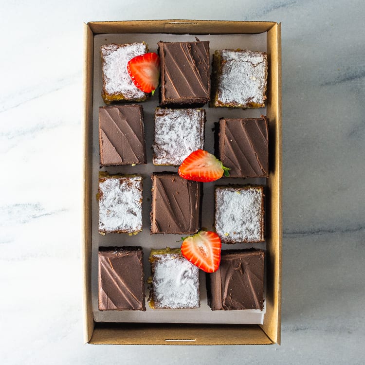 A rectangular cardboard box filled with a mix of 12 sweet slices in orange and almond, and chocolate mud flavours, garnished with fresh strawberry on a marble table.
