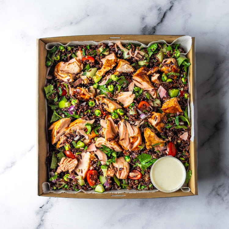 Box of miso-glazed salmon, mixed rice, cherry tomatoes, cucumber, red onion, pickled ginger, coriander, mint and  wasabi peas with a bowl of wasabi mayo on a marble background