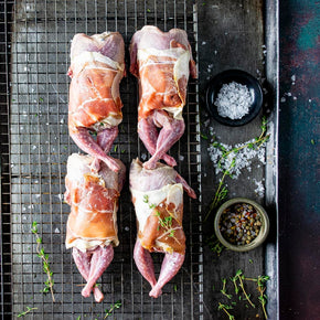 Four mushroom and ricotta stuffed quail wrapped in prosciutto arranged on a wire cooling rack over a baking sheet, with bowls of salt and pepper with herbs nearby.