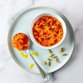 A top-down view of a bowl of Persian Carrot Marmalade and a spoonful of marmalade on a light blue plate, garnished with cardamom seeds, on a white marble background.