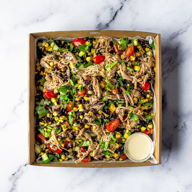 Box with pulled pork, charred corn, black beans, cherry tomatoes, cucumber, red onion, fresh mint, coriander, and a bowl of smoky chipotle cream on a marble background.