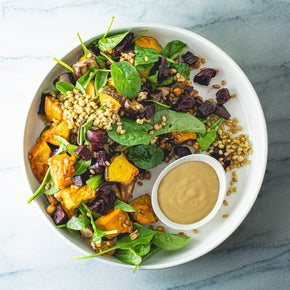 A large round white salad bowl with a mix of roast pumpkin, eggplant, and beetroot, freekeh, spiced chickpeas, and spinach, with a small bowl of balsamic vinaigrette on a marble table.
