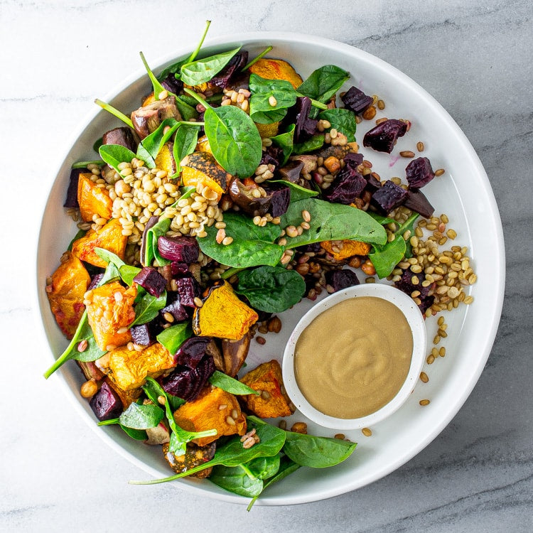 Plated salad with roasted pumpkin, eggplan and beetroot with spinach and a side of tahini dressing on a marble surface.