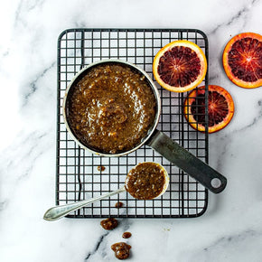 A top-down view of a small metal saucepan filled with Quince, Orange, Seeded Mustard & Ginger Glaze, set on a black wire cooling rack over a white marble surface. A teaspoonful of the sauce sits below the pan, and three sliced blood oranges are arranged on the right side.