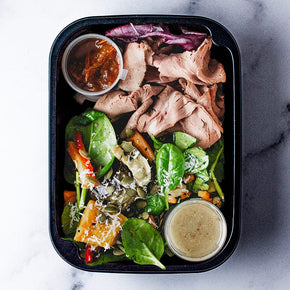 A rectangular container with slices of rare roast beef, salad of pumpkin, eggplant, beetroot, freekeh, chickpeas, and spinach with dressing.