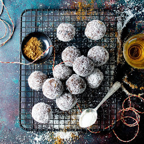 A top-down styled food photo showing several rum balls dusted with coconut on a black wire cooling rack, with a small bowl of spices, a silver teaspoon, and a glass of rum on a dark textured surface.