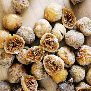 Dried figs, including whole and halved pieces showing their brown seeds, piled on a light wooden surface.