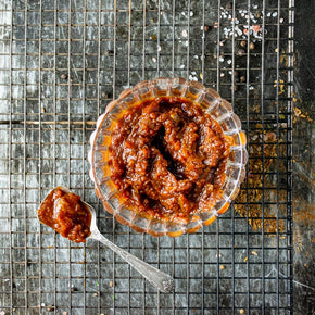 A top-down view of Smoked Spiced Tomato Relish in a small clear glass bowl, with a teaspoon holding a dollop of the relish beside it, set on a dark metal cooling rack.