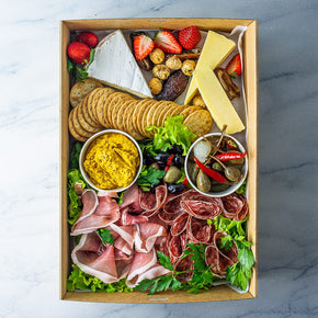A rectangular cardboard box filled with slices of salami, prosciutto, and cheese, with tubs of dip, olives and pickled vegetables, along with crackers, fresh and dried fruit on a marble table.