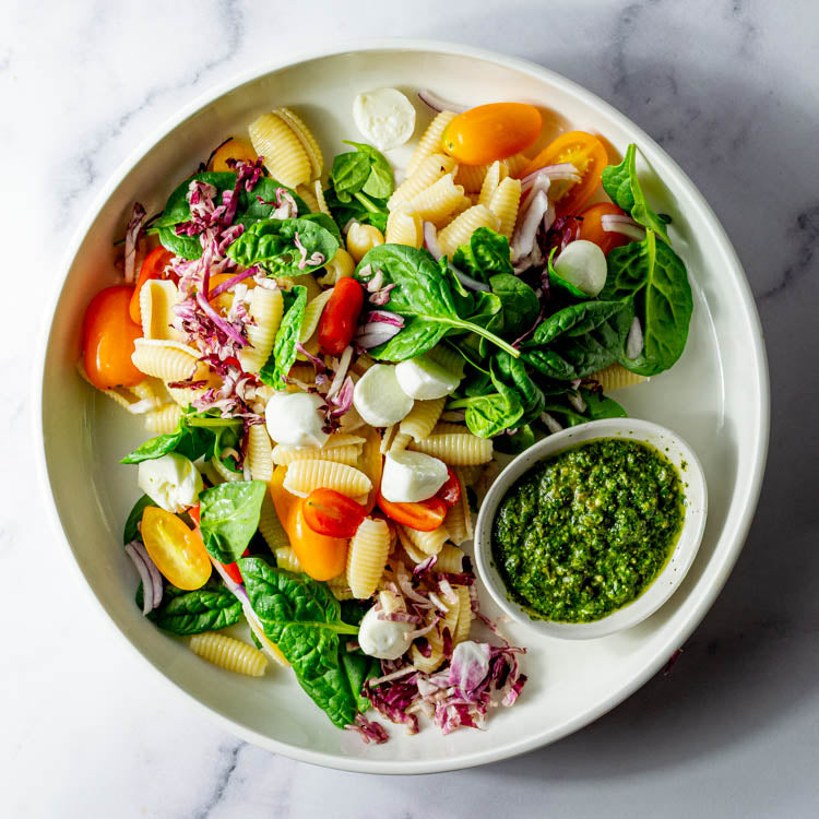 A round white platter filled with a salad of pasta, cherry tomatoes, bocconcini, radicchio, red onion, and basil with small bowl of pesto dressing.