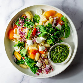 A round white platter filled with a salad of pasta, cherry tomatoes, bocconcini, radicchio, red onion, and basil with small bowl of pesto dressing.