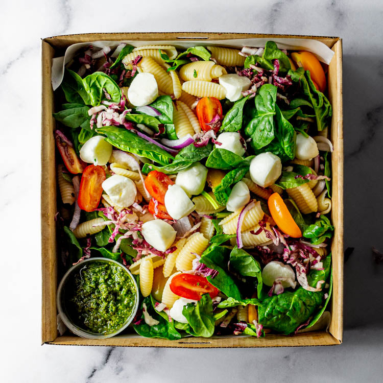 Box filled with a salad of pasta, cherry tomatoes, bocconcini, radicchio, red onion, and basil with tub of pesto dressing on a marble surface