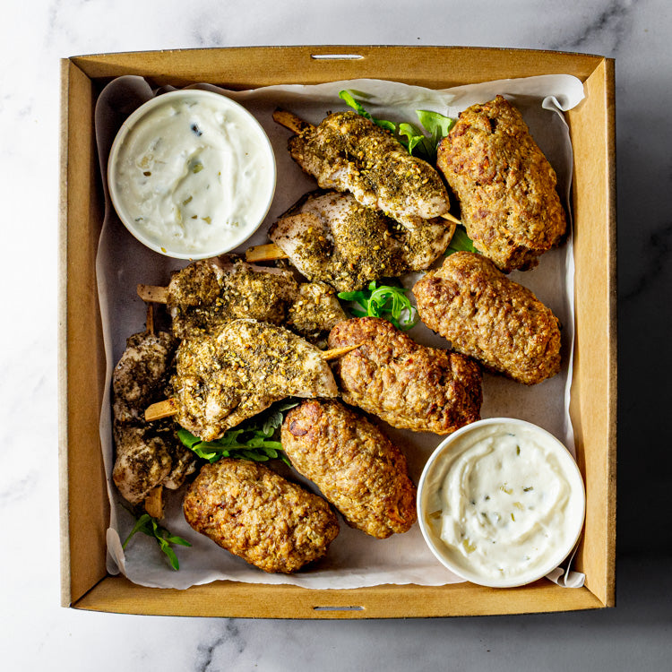 Box of za’atar chicken skewers, lamb kofta and tzatziki on a marble surface.