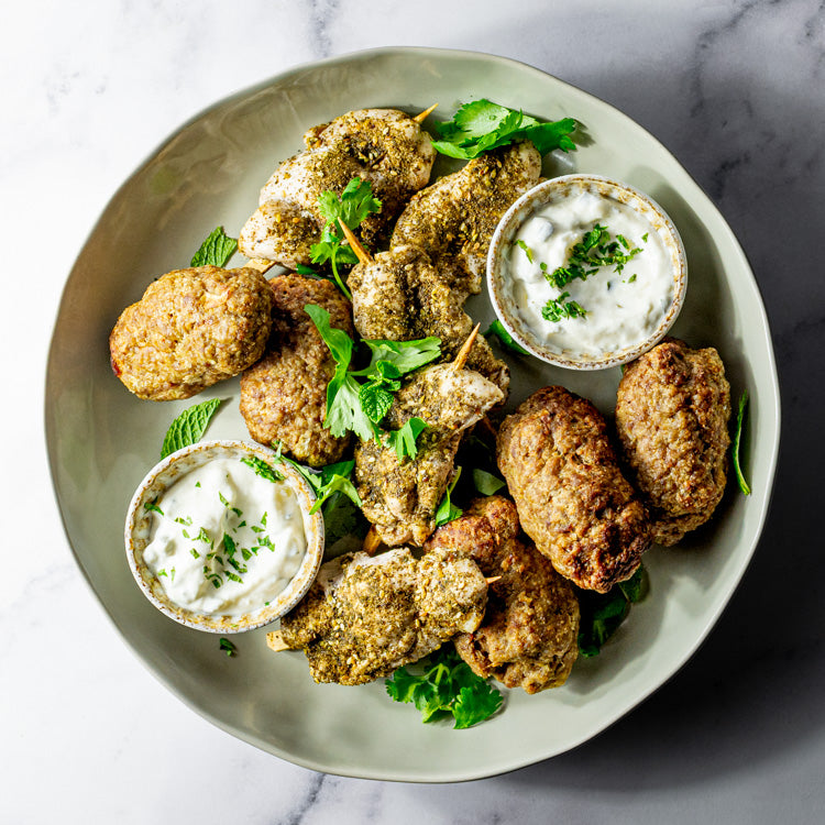 Plate of za’atar chicken skewers, lamb kofta and tzatziki on a marble surface.