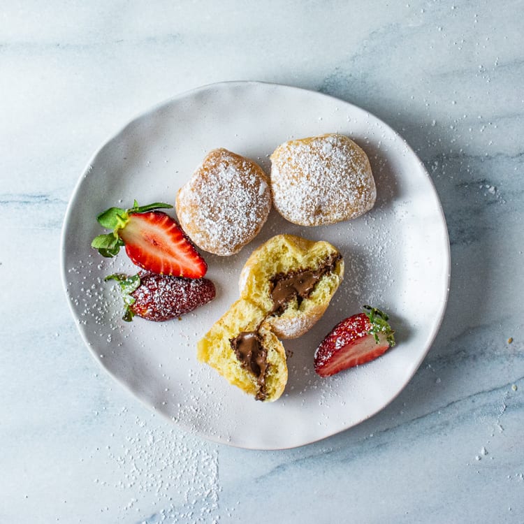 A small white plate with three donuts and strawberry garnish, dusted with icing sugar on a marble table.
