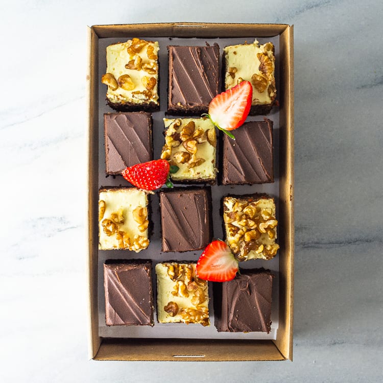 A rectangular cardboard box filled with a mix of 12 sweet slices in carrot and walnut, and chocolate mud flavours, garnished with fresh strawberry on a marble table.