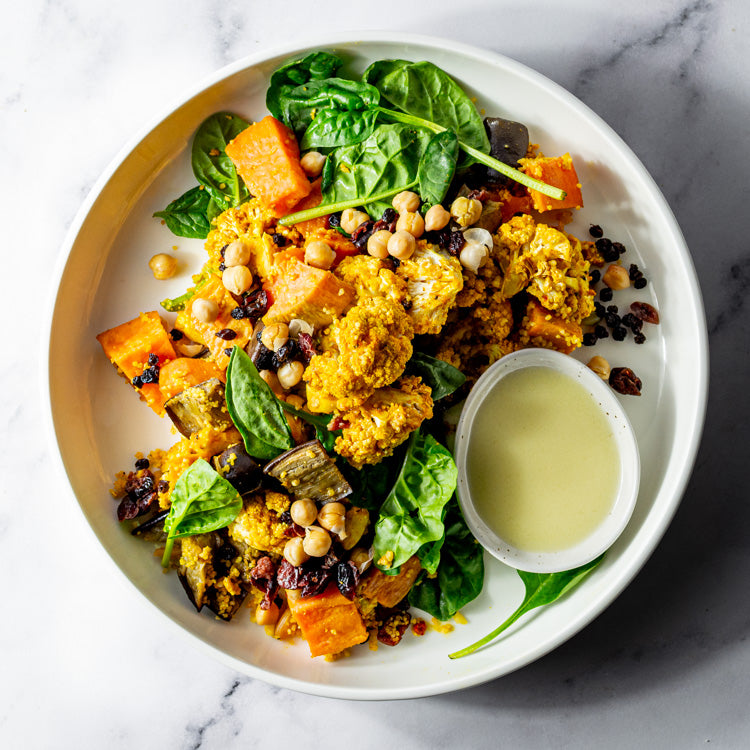 Platter of Moroccan spiced cauliflower, sweet potato, eggplant, chickpeas, baby spinach, cranberries, and currants with bowl of lemon saffron vinaigrette on a marble surface