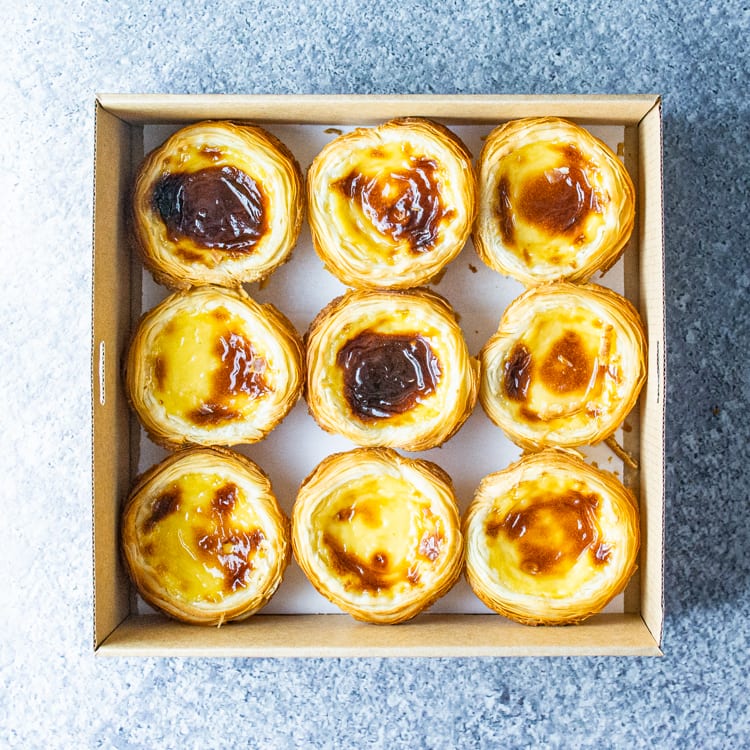 A square cardboard box with nine Portuguese custard tarts on a marble table.