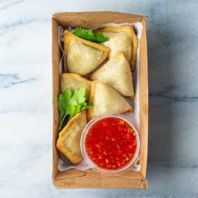 A small rectangular cardboard box containing six golden mini empanadas, tub of sweet chilli sauce, and parsley garnish, on a marble table.