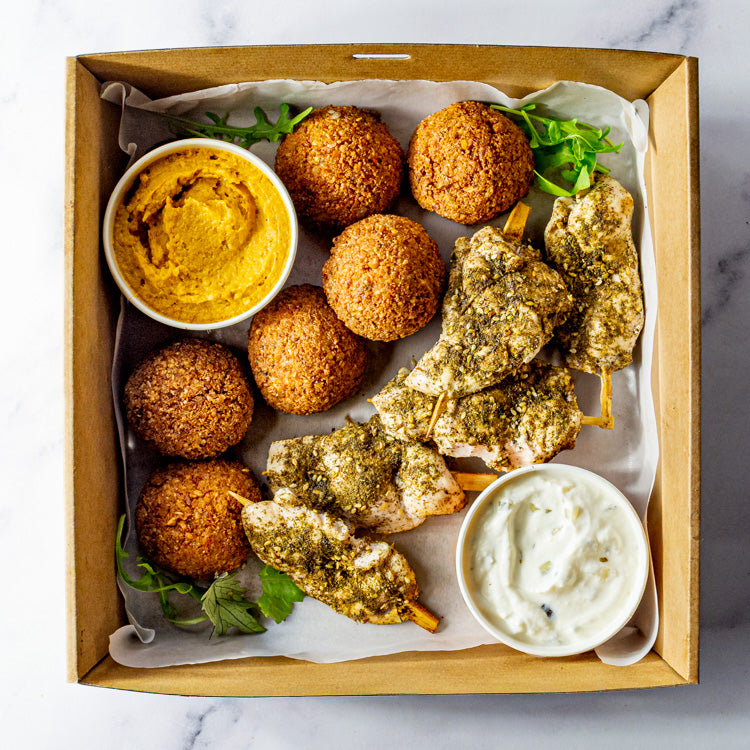 Box of za’atar chicken skewers, falafel, pumpkin hummus and tzatziki on a marble surface