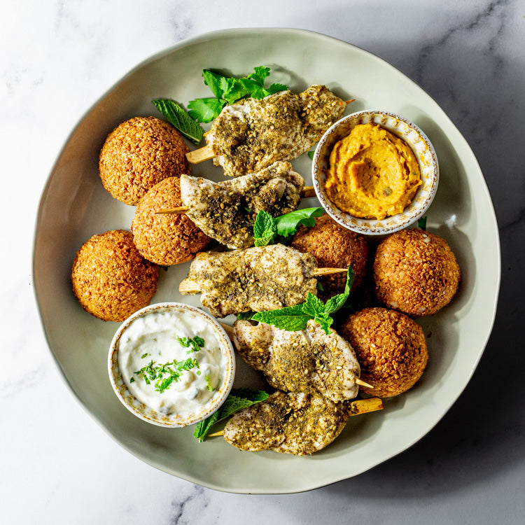 Platter of za’atar chicken skewers, falafel, pumpkin hummus and tzatziki on a marble surface