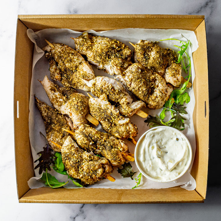 Box of za’atar chicken skewers and tzatziki on a marble surface.
