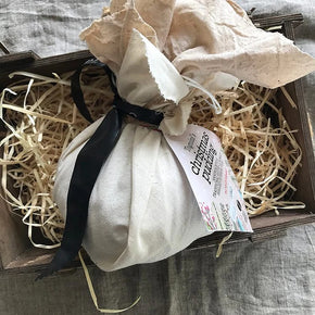 A Traditional Boiled Cloth Wrapped Plum Pudding wrapped in calico and tied with black ribbon resting on a wooden box filled with shredded paper on a gray fabric surface.