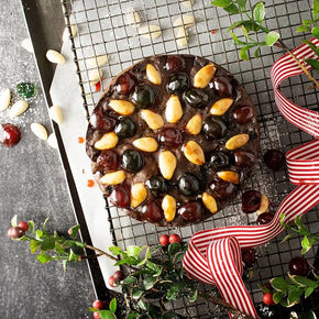 A round Traditional Christmas Cake topped with glossy dark cherries and sliced almonds, presented on a wire cooling rack with holiday decorations including a red-and-white striped ribbon, holly leaves, and scattered cherries.