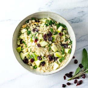 A top-down view of a gluten free Apple & Cranberry Stuffing in a light gray ceramic bowl, set on a white marble surface with fresh sage and cranberries nearby.