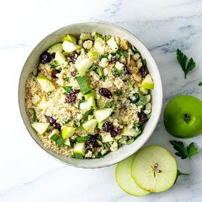 A top-down view of a Apple & Cranberry Stuffing in a light gray ceramic bowl, set on a white marble surface with fresh sage and cranberries nearby.