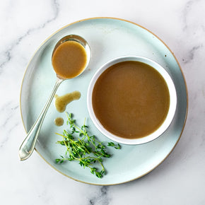 A top-down view of a Turkey Gravy served in a white bowl on a pale blue ceramic plate, with a spoon of gravy, and fresh green herbs on a white marble surface.