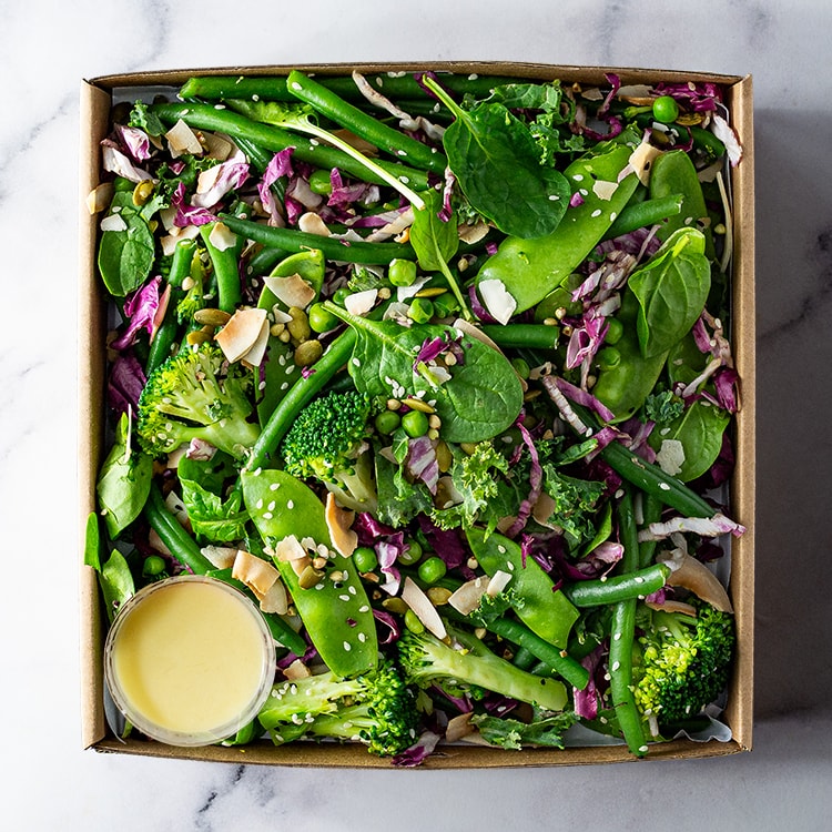 Box of fresh salad with greens, broccoli, and a small container of dressing on a marble surface.