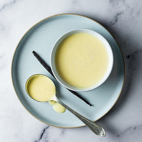 A top-down view of a light blue plate on a white marble surface, holding a white bowl filled with Vanilla Bean Anglaise, alongside one spoon and a vanilla pod.