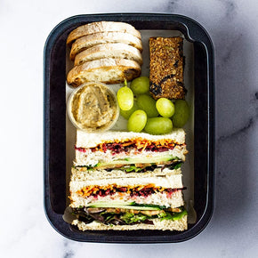 A small rectangular container with two finger sandwiches, crustini, dip, fresh fruit & nut slice on a marble table.