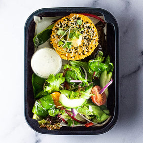 A small rectangular container with a vegan pattie, garden salad, cashew aioli and balsamic dressing on a marble table.
