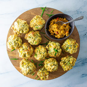 A dozen vegan mini savoury muffins on a round wooden board with a bowl and spoon of relish on the side.