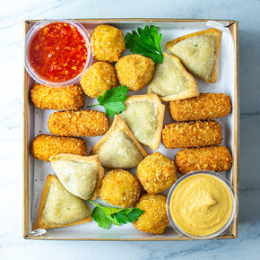 A square rectangular cardboard box containing empanadas, pies, and arancini, with tubs of dipping sauces on a marble table.