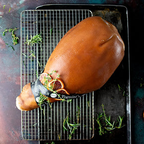 A whole leg of ham, rests on a wire cooling rack over a dark baking sheet. The ham is garnished with dried orange slices, fresh rosemary, and thyme.