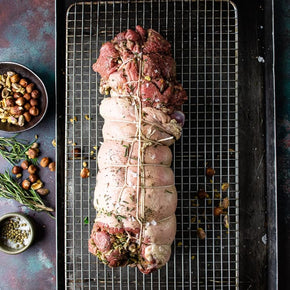 A raw, tied whole rolled deboned lamb leg stuffed with hazelnuts, pistachios and mixed herbs rests on a metal wire rack set over a dark baking sheet. Roasted nuts, rosemary sprigs, and small bowls of seasoning are arranged around it on a dark rustic surface.