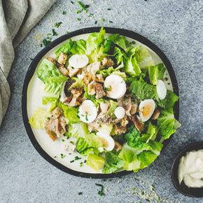 A large round plate bowl with a mix of cos hearts, sourdough croutons, bacon, and boiled eggs, with a small bowl of parmesan aioli dressing on a marble table.