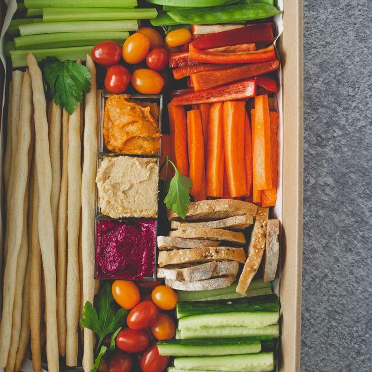 Close up of a rectangular box with a clear lid containing three dips, batons of celery, carrot, cucumber, red and green pepper, cherry tomatoes, crostini, and grissini sticks.
