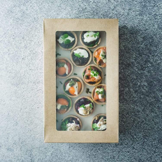 A rectangular cardboard box with a clear lid containing a mix of mini savoury tarts including flavours like hot smoked salmon, beetroot, cherry tomato and caramelised onion, on a marble table.