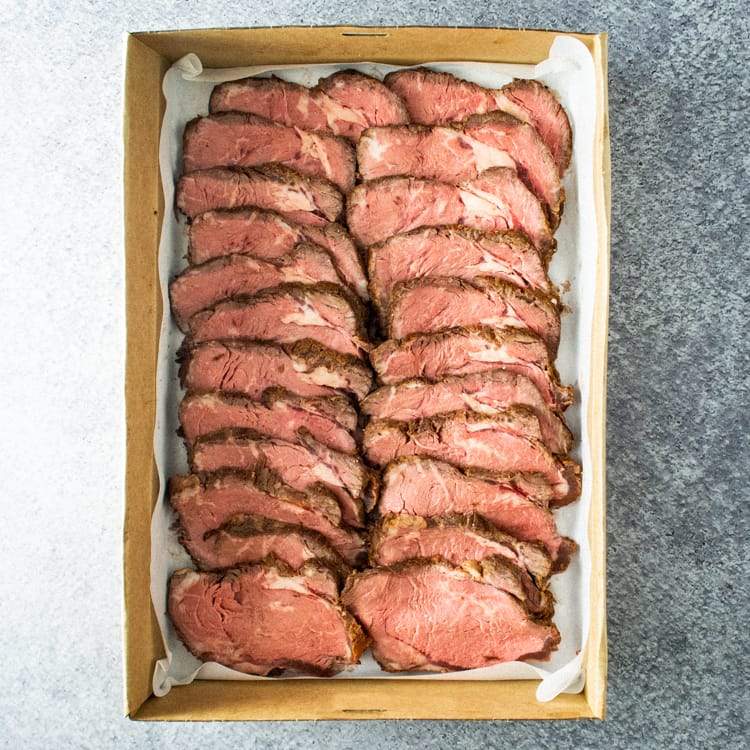 A rectangular cardboard box containing slices of seasoned rib eye fillet, on a marble table.