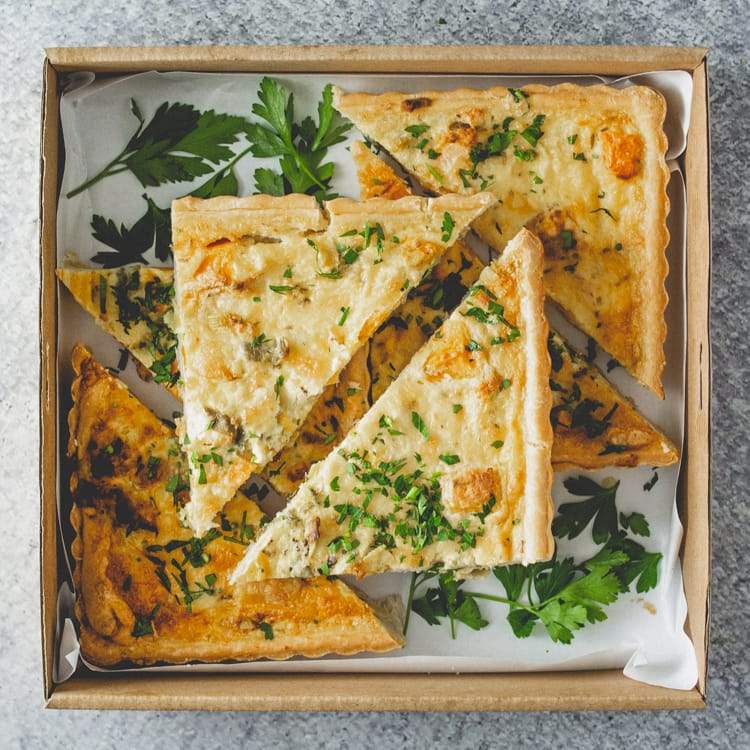 A square cardboard box containing triangular slices of roast pumpkin, leek, and feta tart, garnished with parsley, on a marble table.