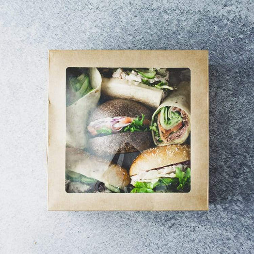 A square cardboard box with a clear lid containing an assortment of rolls and wraps with a mix of fillings, on a marble table.