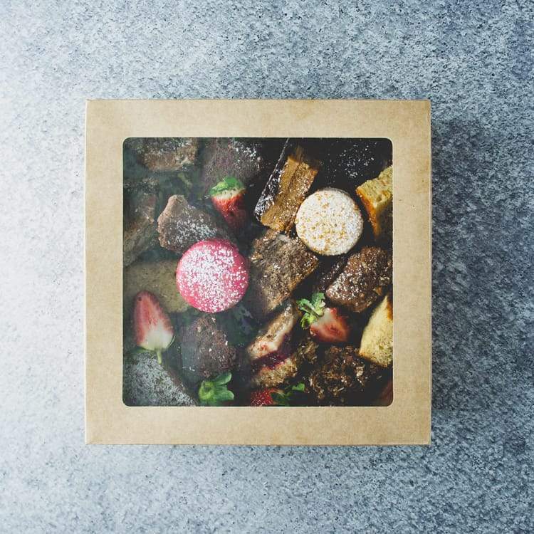 A square cardboard box with a transparent lid filled with assorted sweet slices and macarons, dusted in icing sugar with a strawberry garnish on a marble table.